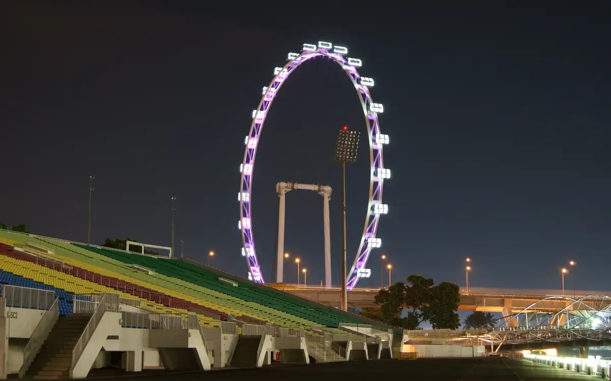 The Singapore Flyer lit up at night beside a sports stadium grandstand, with the wheel’s white lights against a dark sky